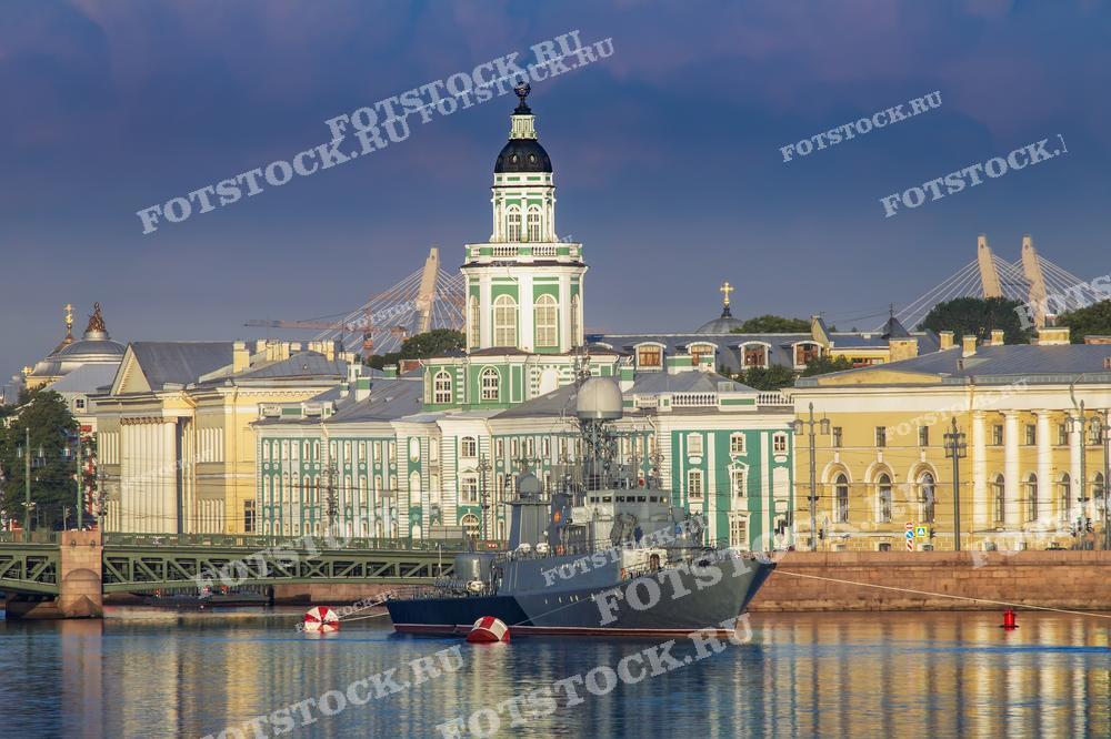 На фотографии запечатлен утренний или вечерний вид на Кунсткамеру и часть Университетской набережной Санкт-Петербурга. Воды Невы отражают здания и мягкий свет, добавляя кадру тишины и спокойствия. Виден также фрагмент моста и корабль на переднем плане, который вносит в композицию элемент морской тематики. Изображение подходит для использования в туристических гидах, культурно-исторических изданиях, а также как элемент декора.